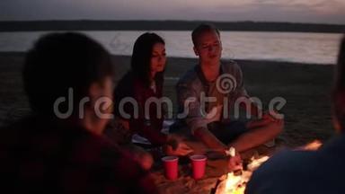 可爱的夫妇在篝火上烤棉花糖，接吻。 年轻人坐在海滩上的<strong>朋友圈</strong>里